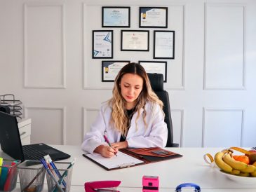 Nutritionist sits at a modern office desk writing, with fruits and diplomas displayed, emphasizing health and professionalism.
