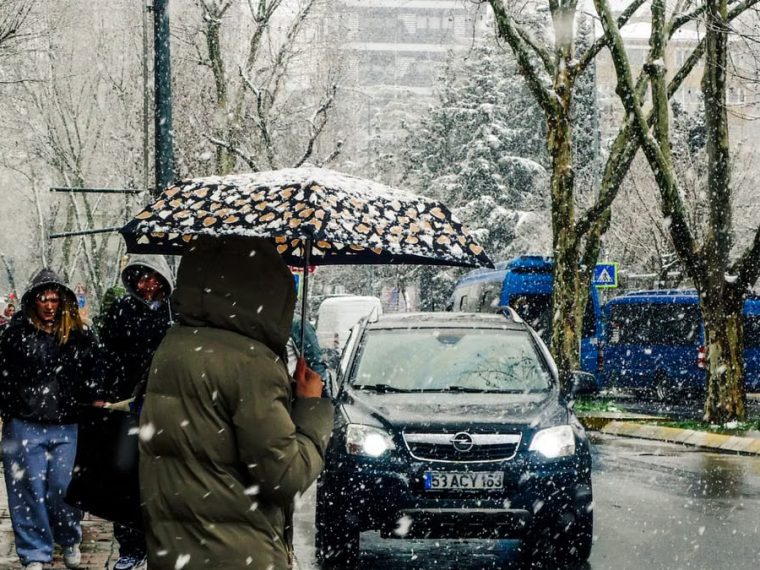 A snowy city street with people carrying umbrellas and cars passing by.